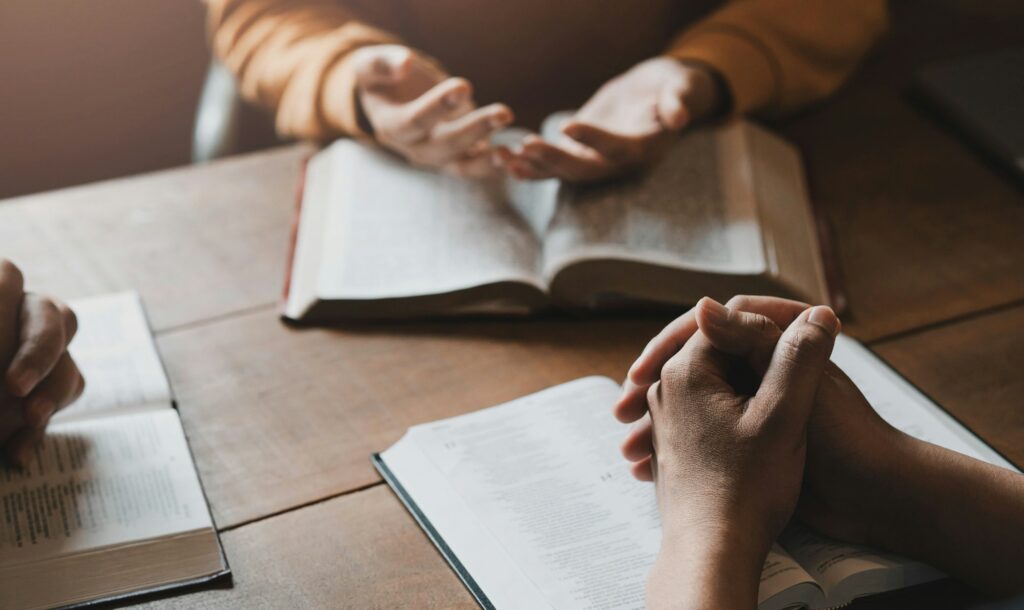 A group of Christians sit together and pray around a wooden table with blurred open Bible pages